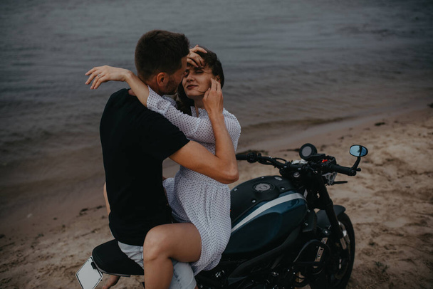 Couple in love hugs on beach sitting on motorcycle on water background. - Photo, Image