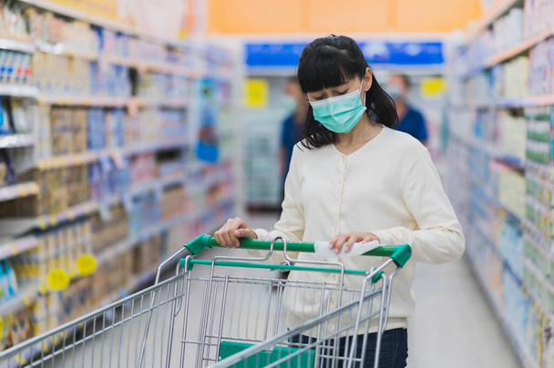 Азиатка, носящая медицинскую маску для лица, стирает ручку тележки в супермаркете, чтобы предотвратить распространение микробов и бактерий и избежать инфекций коронного вируса. Концепция гигиены - Фото, изображение