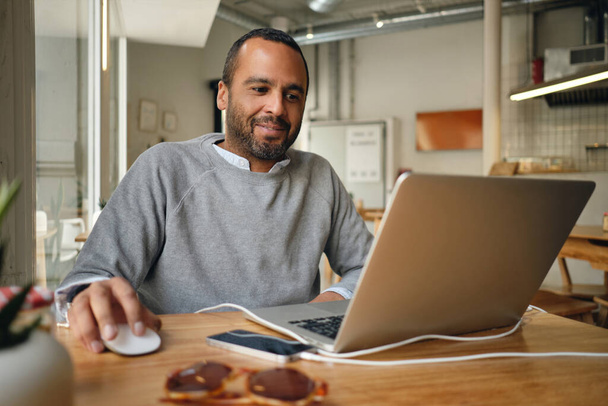 Uomo d'affari in maglione felicemente lavorando sul computer portatile durante la pausa caffè in città caffè
 - Foto, immagini