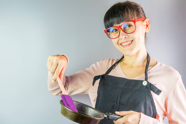 Ασιάτισσα γυναίκα διασκεδάστε νοικοκυρά φορώντας ποδιά κουζίνας μαγείρεμα με τηγάνι. - Φωτογραφία, εικόνα
