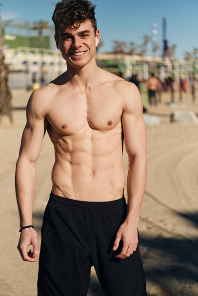 Joven hombre deportivo alegre con el torso desnudo felizmente mirando en la cámara después del entrenamiento al aire libre
 - Foto, Imagen