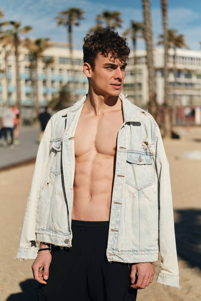 Joven chico deportivo con confianza en chaqueta de mezclilla con el torso desnudo posando en la cámara en la playa de la ciudad
 - Foto, Imagen