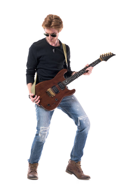 引き裂かれたジーンズの若いスタイリッシュなハンサムな赤い髪のロッカーは、電気ギターを見下ろしています。白い背景に隔離された全身の長さ.  - 写真・画像