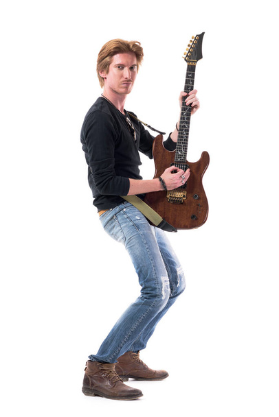 Πλευρική άποψη της αυτοπεποίθηση ροκάς τζίντζερ παίζει ηλεκτρική κιθάρα κοιτάζοντας κάμερα. Πλήρες μήκος σώματος που απομονώνεται σε λευκό φόντο.  - Φωτογραφία, εικόνα