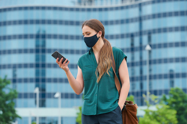 Νεαρή γυναίκα με μάσκα προσώπου για να αποφύγει την εξάπλωση του κορωναϊού στο κέντρο της πόλης. Κορίτσι με μακριά μαλλιά διαβάζοντας τις ειδήσεις στο smartphone σπρώχνοντας ένα χέρι σε μια τσέπη του παντελονιού στο κέντρο - Φωτογραφία, εικόνα