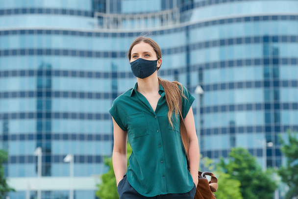 A pretty young woman in a medical face mask to avoid the spread of coronavirus walking in the center of the city. A girl with long hair keeping social distance wears a protective face mask in downtown - Photo, Image