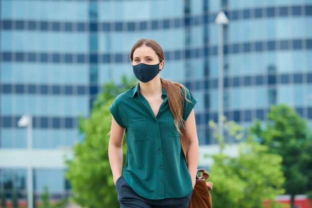 Una mujer con una máscara facial médica para evitar la propagación del coronavirus caminando por el centro de la ciudad. Una chica con el pelo largo manteniendo la distancia social lleva una máscara protectora en el centro de la ciudad
. - Foto, imagen