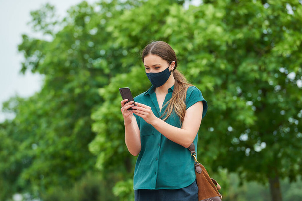 Молодая женщина в синей морской маске для лица читает новости на смартфоне во время прогулки по парку. Девушка в защитной маске держит социальную дистанцию, чтобы избежать распространения коронавируса рядом с деревьями
. - Фото, изображение