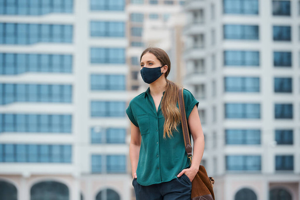 Tıbbi maske takan güzel bir kadın, şehrin ortasında yürürken ellerini pantolon ceplerine sokuyor. Sokakta sosyal mesafeyi koruyan koruyucu maskeli bir kız.. - Fotoğraf, Görsel