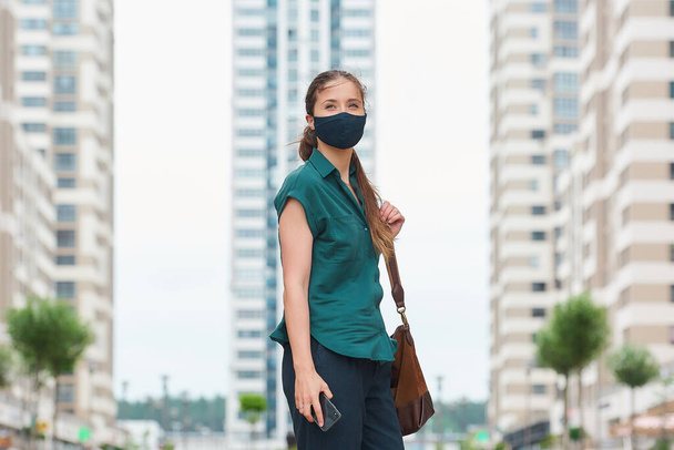 スマートフォンを持っている医療用フェイスマスクの若い女性が別の手をズボンのポケットに投げ込み、高層ビルの間を歩く。保護顔のマスクをしたまま社会的距離を保つ少女. - 写真・画像
