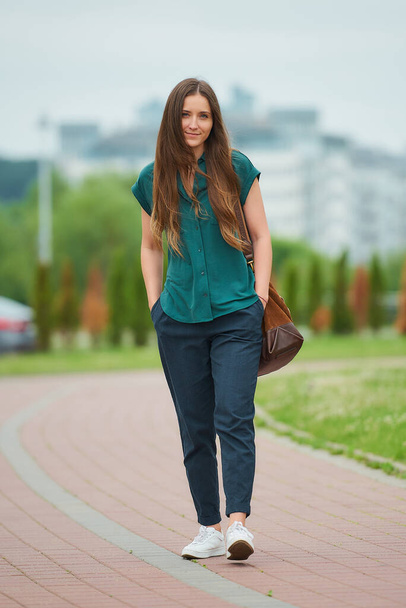 Молодая женщина в стильной повседневной одежде ходит, засовывая руки в карманы брюк в парке. Красивая девушка носит офисную одежду, сохраняя социальную дистанцию в городе
.  - Фото, изображение