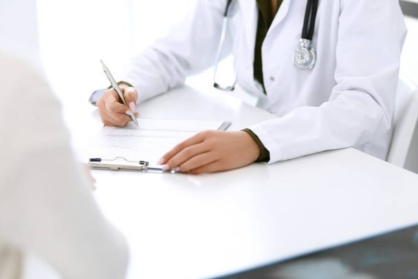 Woman doctor and patient sitting and talking at medical examination at hospital office, close-up. Therapist filling up medication history records. Medicine and healthcare concept - Photo, image