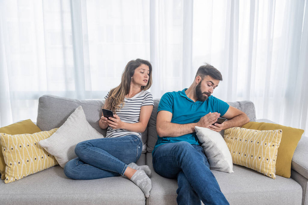 Young boyfriend making fun and jealous his girlfriend pretending that he is texting with other girl on his cell or mobile phone while they sitting on the sofa in their apartment - Foto, Bild