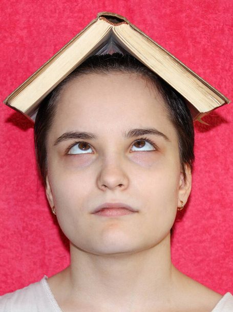 Πορτρέτο ενός κοριτσιού με ένα βιβλίο στο κεφάλι της σε σχήμα στέγης. Εκμάθηση εξ αποστάσεως. - Φωτογραφία, εικόνα