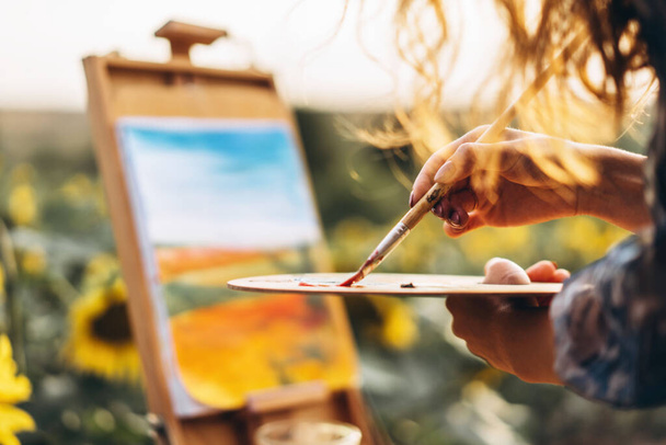 油絵具でブラシやパレットを保持する女性アーティストの手を閉じます。ひまわり畑でのイーゼルの背景が赤く. - 写真・画像