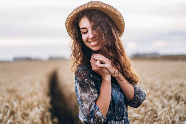 Closeup portrait of a beautiful young woman with curly hair. Woman in dress and hat standing in wheat field. - Фото, изображение