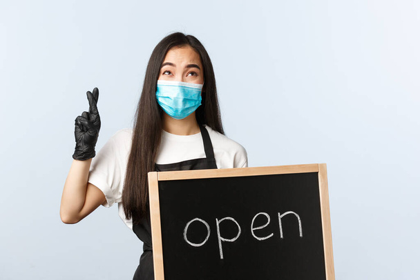 Small business, covid-19 pandemic, preventing virus and employees concept. Hopeful young asian cafe owner, store worker in medical mask cross fingers, praying as holding open sign - Foto, imagen