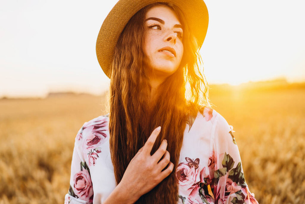 Portrait of a beautiful young woman with curly hair and freckles face. Woman in dress and hat posing in wheat field at sunset and looking at camera. - Φωτογραφία, εικόνα