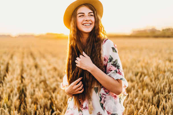 Portrait of a beautiful young woman with curly hair and freckles face. Woman in dress and hat posing in wheat field at sunset and looking at camera. - Foto, imagen