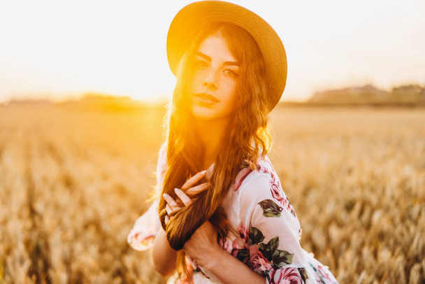 Portrait of a beautiful young woman with curly hair and freckles face. Woman in dress and hat posing in wheat field at sunset and looking at camera. - Фото, изображение