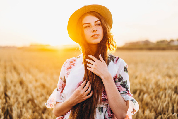 Portrait of a beautiful young woman with curly hair and freckles face. Woman in dress and hat posing in wheat field at sunset and looking at camera. - Foto, afbeelding