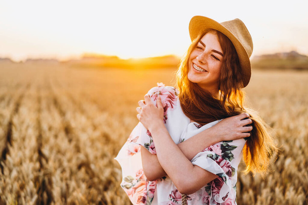 Incredible young woman with long curly hair and freckles face. Woman in dress posing in wheat field at sunset. Close up portrait. - Foto, afbeelding