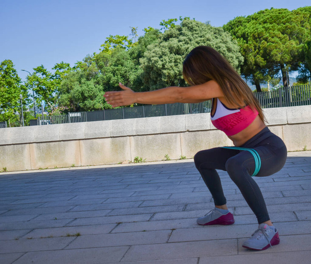 Girl training outdoors, doing squats with elastic bands in the park - Photo, Image