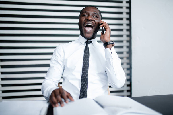 Portrait of a black african american businessman in a white shirt sitting at a table and talking emotionally on the phone with his mouth out loud. - Fotó, kép