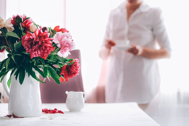 Woman having cup of coffee in the kitchen decorated with pink peonies. - Photo, Image