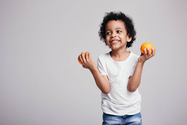 Dark-skinned afro-american curly boy in a white T-shirt and jeans holds two orange oranges in the studio on white background - Photo, image