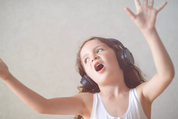 excited small caucasian pretty little girl in black headphones singing song waving hands indoors - Fotó, kép