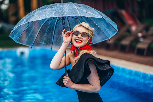 Pin up styled blonde in sunglasses, with red lips and reto hairstyle, in black vintage swimsuite and red scarf, holding umbrella and have fun in the swimming pool - Photo, image
