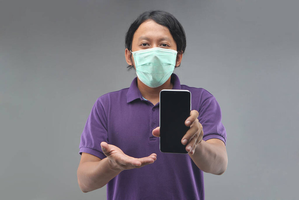 nieuwe normale Indonesië man wijzen smartphone met paars shirt geïsoleerd op grijze achtergrond - Foto, afbeelding