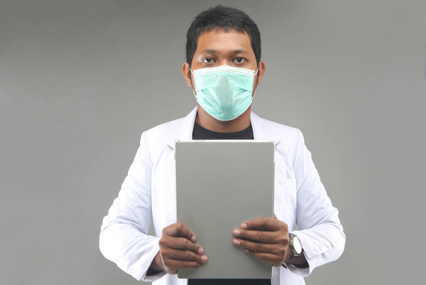 asiatische Mann Portrait Arzt mit Maske oder Gesichtsmaske hält Tabletten Computer isoliert auf grauem Hintergrund. yogyakarta indonesien. 29. September 2020 - Foto, Bild