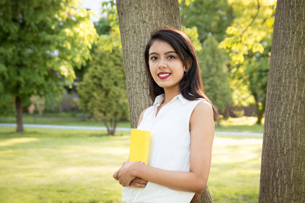 Genç, güzel bir Hintli kız elinde sarı bir defterle bir ağacın yanında duruyor. Açık havada bir kız öğrencinin portresi. Kız dışarıda karantinada vakit geçiriyor. - Fotoğraf, Görsel