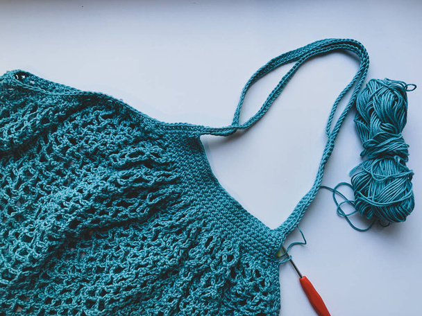 String bag in the process of knitting. Turquoise mesh bag, crochet hook and remaining yarn on a white background. The concept of creating a product. - Фото, изображение