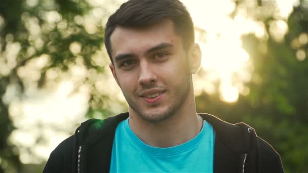 Slow motion of smiling young adult man look at camera in and stand in park in rays of sunset - Footage, Video