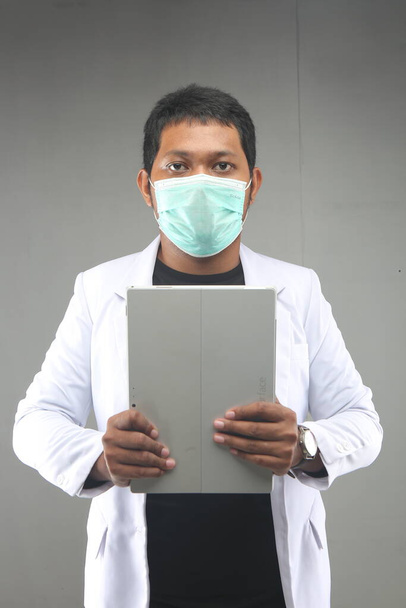 neue normale thailändische Arzt covid-19 mit Gesichtsmaske hält Tabletten Computer isoliert auf grauem Hintergrund. yogyakarta indonesien. 29. September 2020 - Foto, Bild