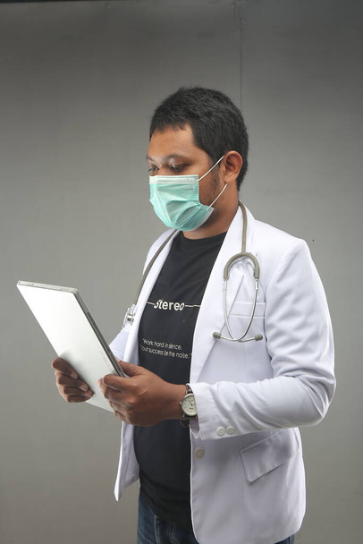 neue normale asiatische männliche Arzt covid-19 mit Gesichtsmaske hält Tabletten Computer isoliert auf grauem Hintergrund. yogyakarta indonesien. 29. September 2020 - Foto, Bild
