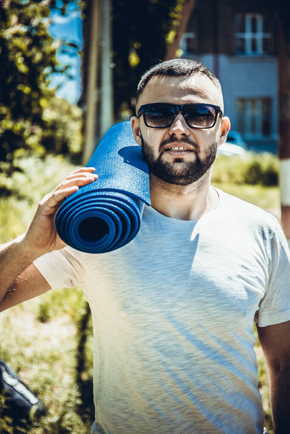 Joven deportista barbudo sosteniendo una alfombra azul en el parque. Hombre joven practicando yoga ejercicio de fitness al aire libre
. - Foto, Imagen