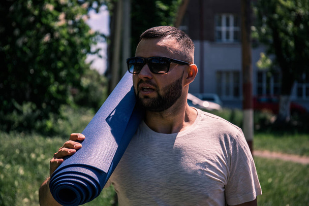 Joven deportista barbudo sosteniendo una alfombra azul en el parque. Hombre joven practicando yoga ejercicio de fitness al aire libre
. - Foto, Imagen