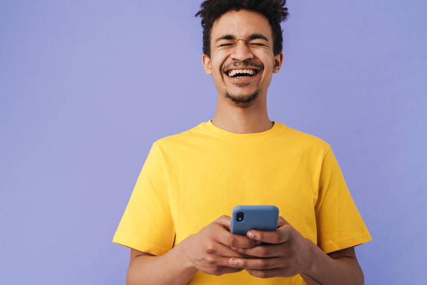 Photo of funny african american man laughing and using smartphone isolated over purple background - Photo, Image