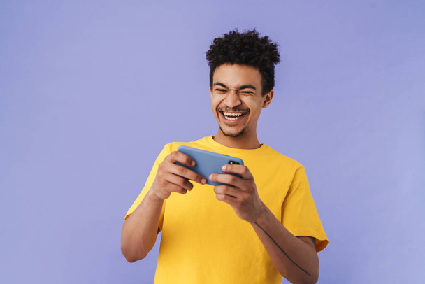 Photo of laughing african american man playing video game on smartphone isolated over purple background - Foto, Bild
