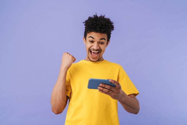 Photo of delighted african american man using smartphone and making winner gesture isolated over purple background - Zdjęcie, obraz