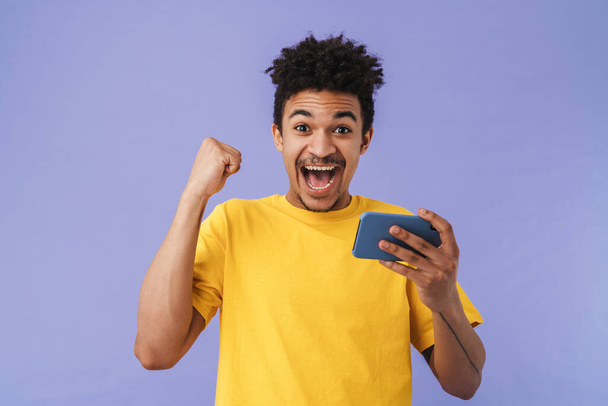 Photo of delighted african american man using smartphone and making winner gesture isolated over purple background - Фото, изображение