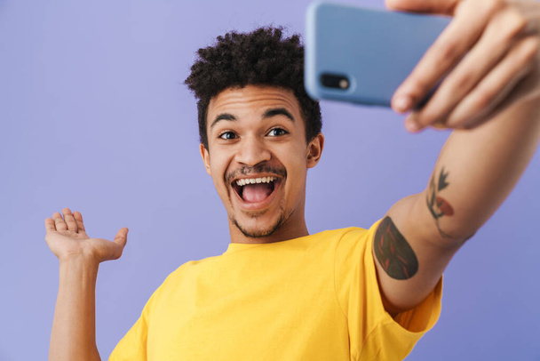 Photo of cheerful african american man taking selfie on smartphone and holding copyspace isolated over purple background - Foto, afbeelding