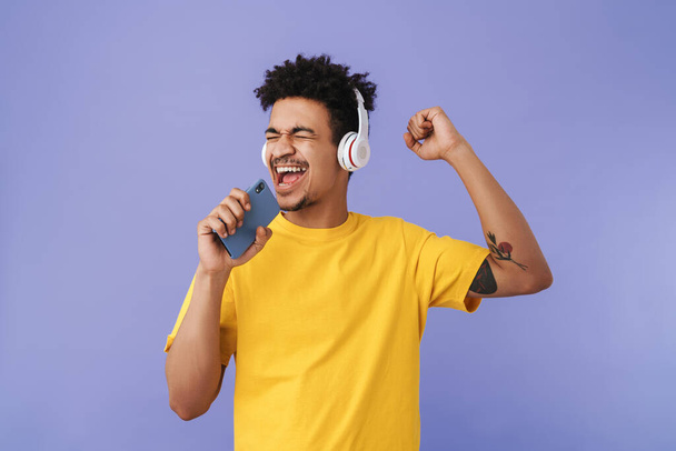 Photo of joyful african american man singing while using cellphone and wireless headphones isolated over purple background - Φωτογραφία, εικόνα