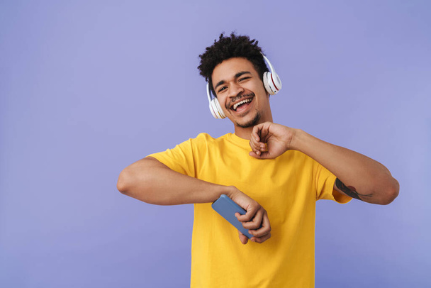 Photo of joyful african american man dancing while using cellphone and wireless headphones isolated over purple background - Foto, Bild