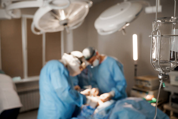 Хирург вставляет силиконовый имплантат в грудь женщины. Увеличение сисек пластическая операция и коррекция в медицинской клинике. Группа профессиональных медиков в медицинских масках и перчатках делают пластическую операцию
 - Фото, изображение