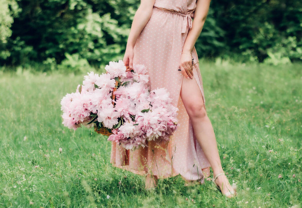 a girl in a pink dress in peas on a green background holds in one hand a basket with pink peonies and in the other glasses with a gold rim - Foto, Imagem